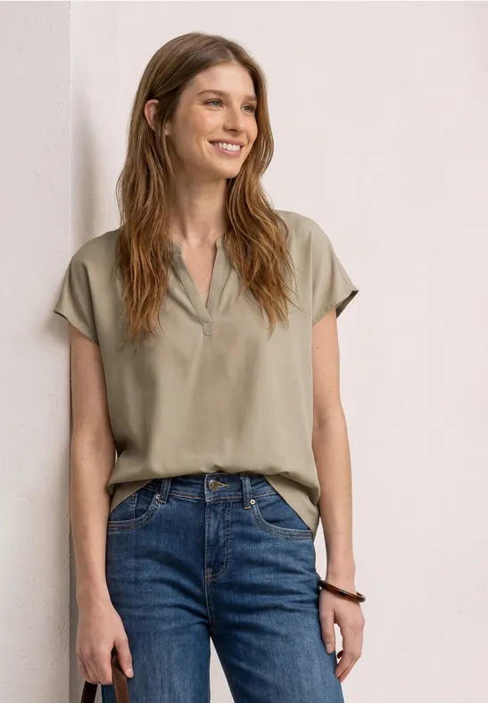 Eine Frau in beiger Bluse und blauen Jeans vor hellem Hintergrund.