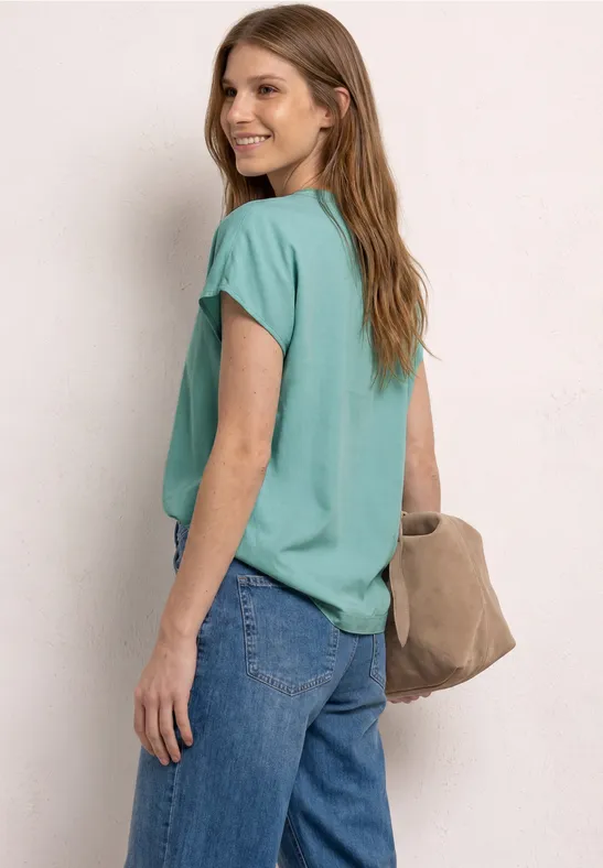 Femme en t-shirt turquoise, jean bleu et sac beige, regard par-dessus l'épaule.