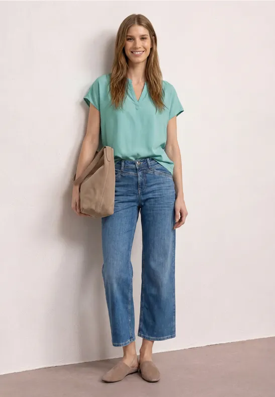 Femme devant un mur clair, porte un T-shirt vert menthe, jean large 7/8 bleu, sac beige et mocassins.
