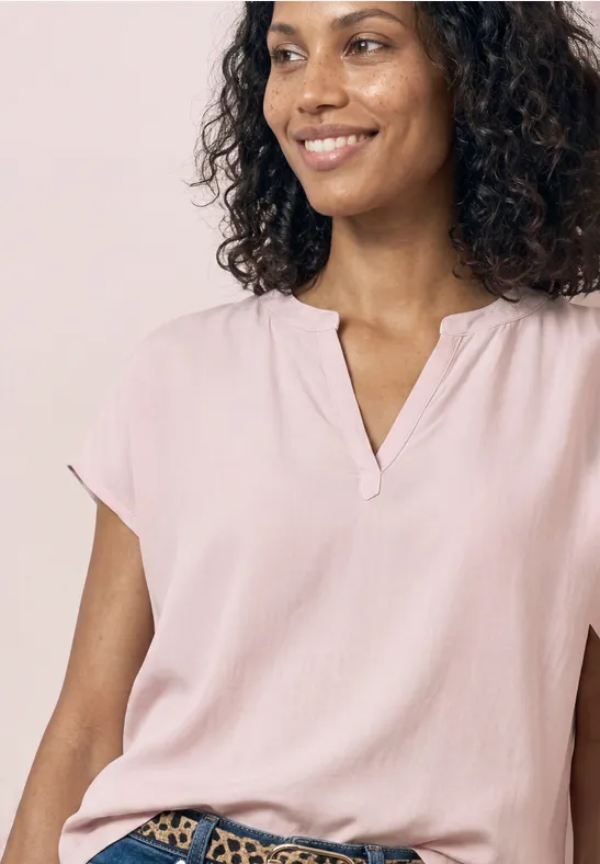 Oberkörper einer Person mit lockigem Haar, rosa Bluse und V-Ausschnitt vor hellem Hintergrund.