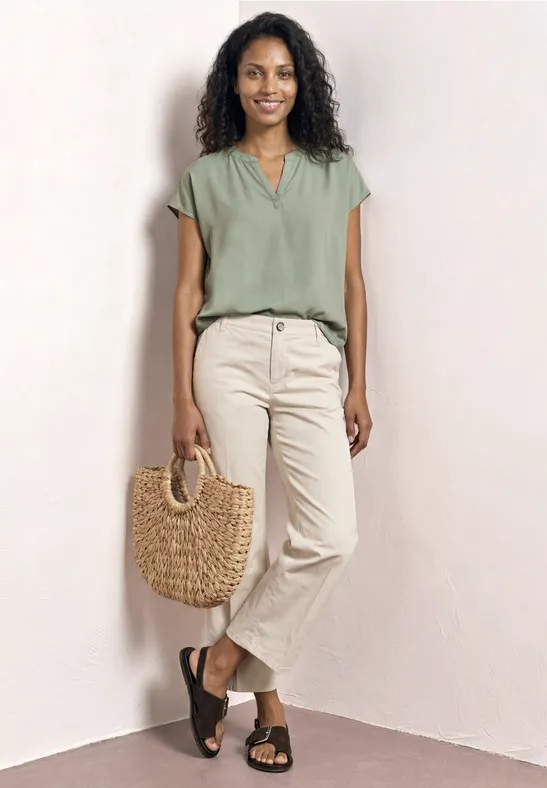 Frau lehnt an hellem Hintergrund, trägt grüne Bluse, helle Hose, schwarze Sandalen und einen geflochtenen Korb.