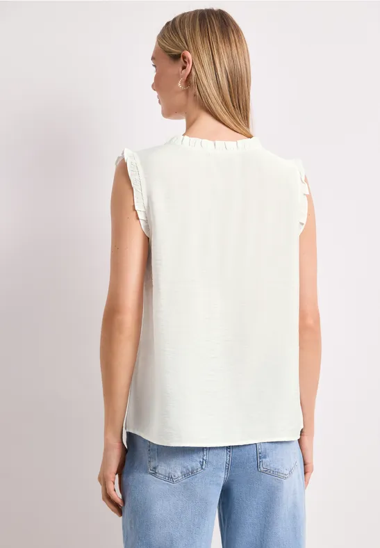 Frau steht seitlich mit dem Rücken zur Kamera, trägt weiße ärmellose Bluse mit Rüschen und blaue Jeans.