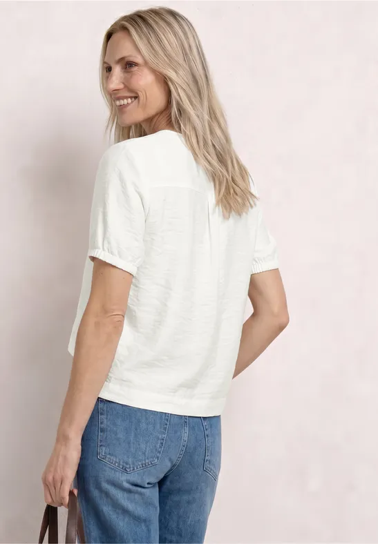 Frau von hinten in weißem T-Shirt und blauen Jeans, hält schwarze Tasche, lockere blonde Haare, neutraler Hintergrund.