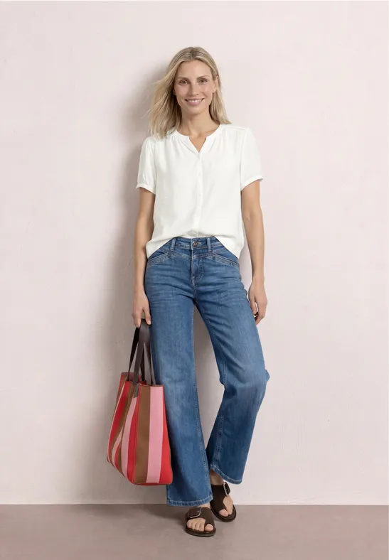 Frau mit weißer Bluse, blauen Jeans und gestreifter Tasche steht vor hellem Hintergrund.