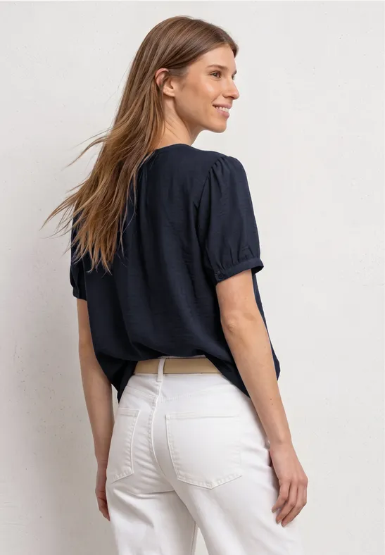 Frau von hinten mit dunkelblauem T-Shirt und weißer High-Waist-Hose vor neutralem Hintergrund.