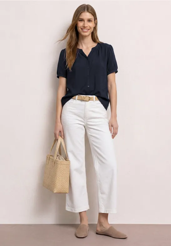 Frau in dunkelblauer Bluse, weißen Culottes, beigen Loafers und Basttasche vor hellem Hintergrund.