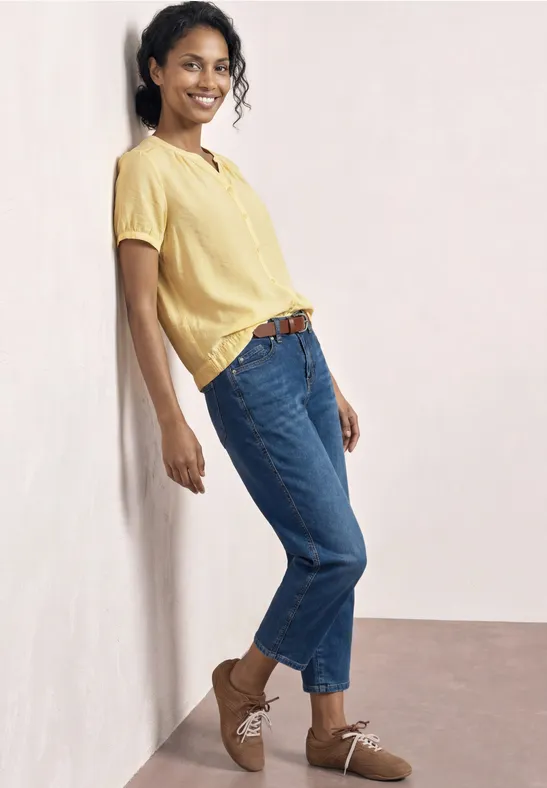 Frau lehnt entspannt in gelbem Shirt, blauen Jeans und braunen Schuhen an heller Wand.