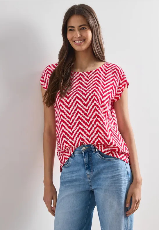 Eine Frau trägt ein rot-weiß gestreiftes Zickzack-Shirt und blaue Jeans vor hellem Hintergrund.