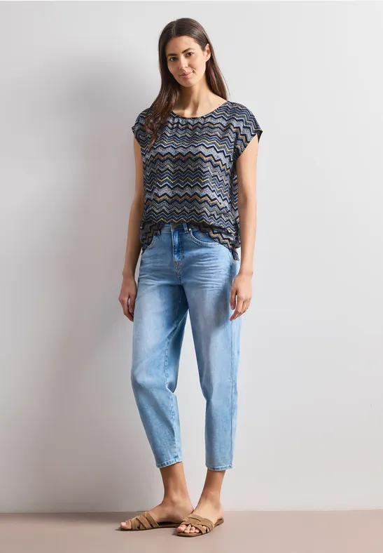 Frau steht vor grauem Hintergrund, trägt gemustertes Oberteil, blaue gekrempelte Jeans und Sandalen.