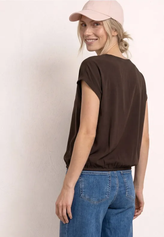 Persoon met bruin T-shirt, blauwe jeans en pet staand zijwaarts voor een lichte achtergrond.