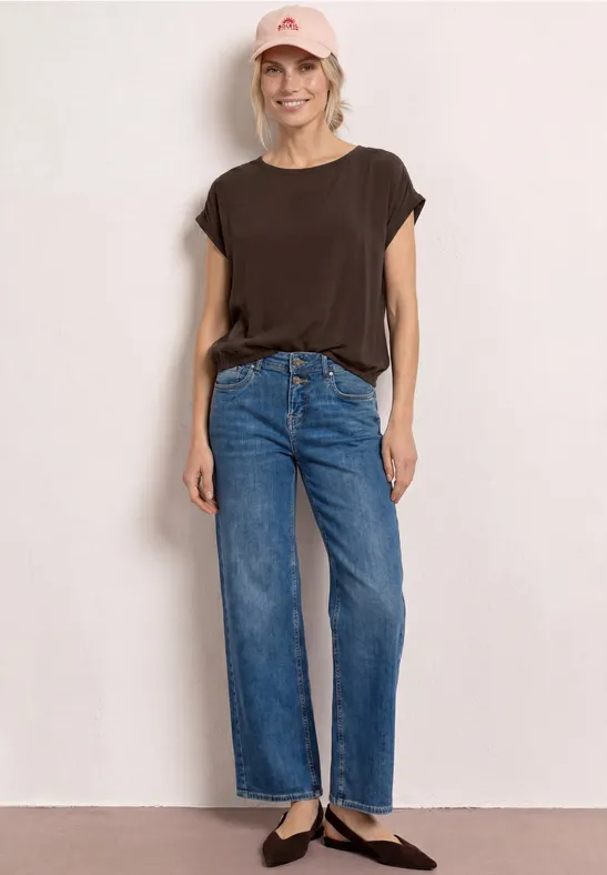 Vrouw staat voor een witte muur, draagt een donkerbruin t-shirt, rechte blauwe jeans en zwarte ballerina’s.