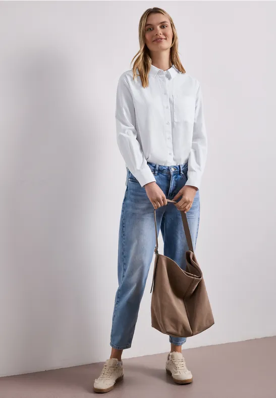 Vrouw draagt witte blouse, wijde blauwe spijkerbroek, beige schoenen en houdt een bruine tas vast.