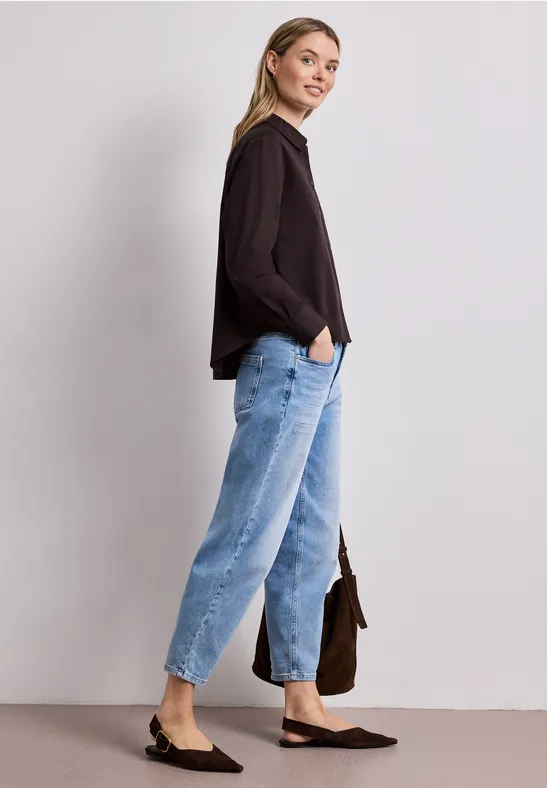 Femme en chemisier noir, jean bleu clair, ballerines pointues noires, de profil avec sac.