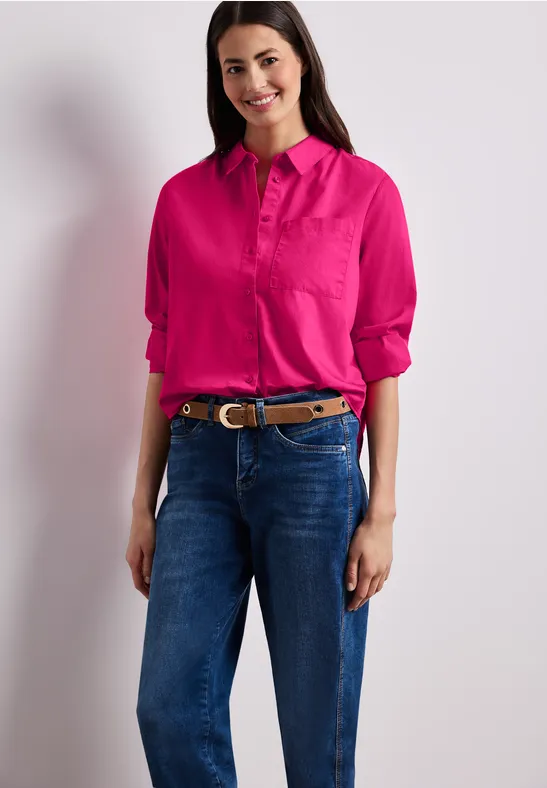 Eine Person trägt ein pinkes Hemd und blaue Jeans vor weißem, neutralem Hintergrund.