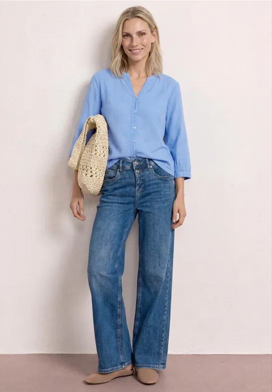 Kobieta w jasnoniebieskiej koszuli, szerokich dżinsach i plecionej słomkowej torbie na jasnym tle.