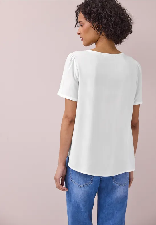 Vrouw van achteren in een los wit t-shirt en blauwe jeans tegen een neutrale achtergrond.