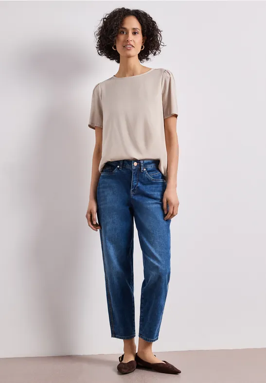 Person in beigem T-Shirt, blauen Jeans und braunen Loafers steht vor weißem Hintergrund.