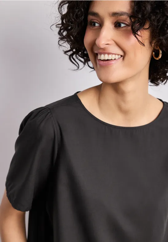 Gros plan d'une personne vêtue d'un chemisier noir à encolure ronde, manches courtes et boucle d'oreille dorée.