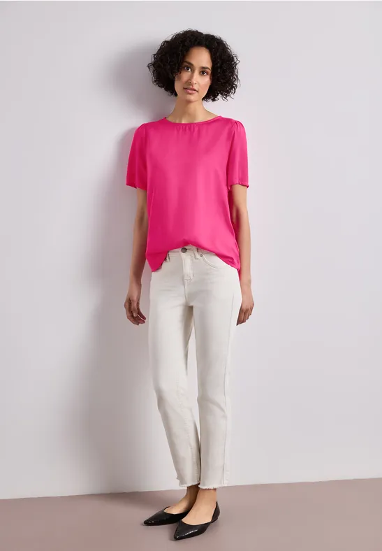 Frau in pinkem T-Shirt und weißer Hose vor hellem Hintergrund.