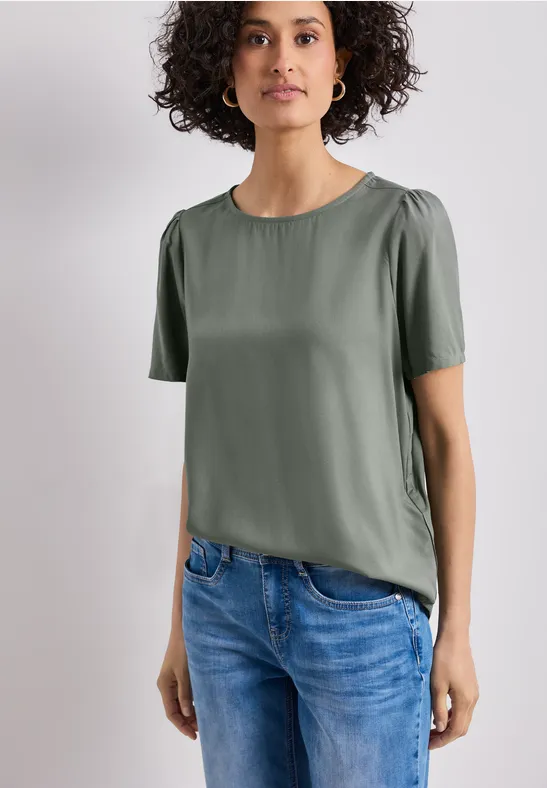 Person trägt grüne Bluse mit kurzen Ärmeln und blaue Jeans vor hellem Hintergrund.