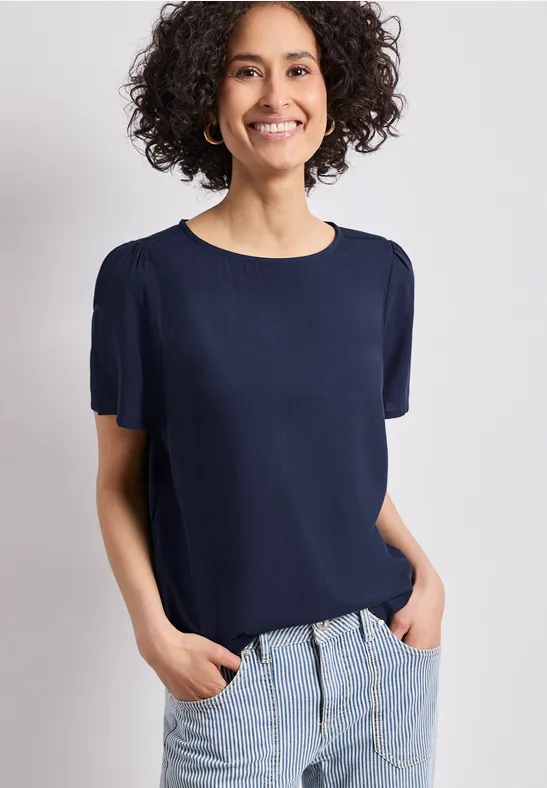 Vrouw staat met handen in haar zakken, draagt een marineblauw shirt met korte mouwen en een lichte spijkerbroek.