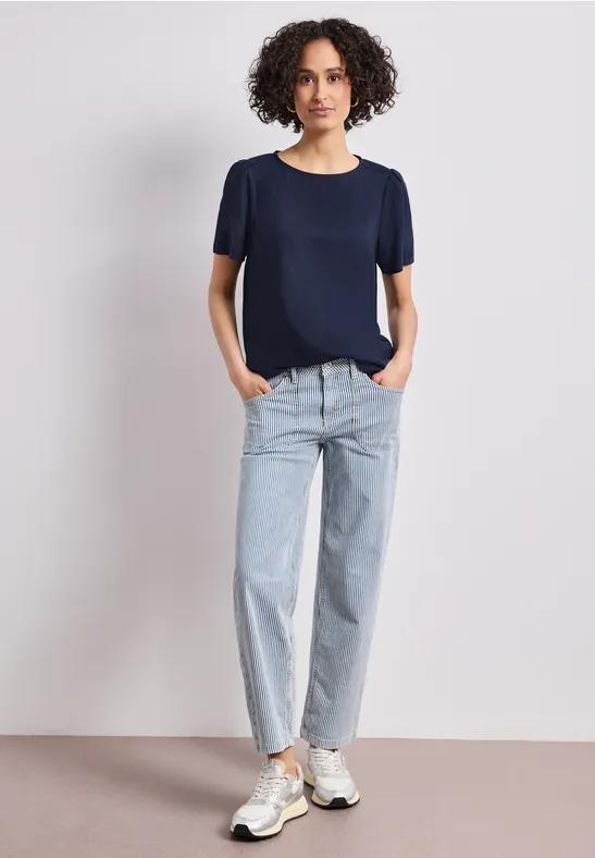 Een vrouw in een donkerblauw t-shirt, lichte jeans en sneakers staat ontspannen tegen een witte achtergrond.