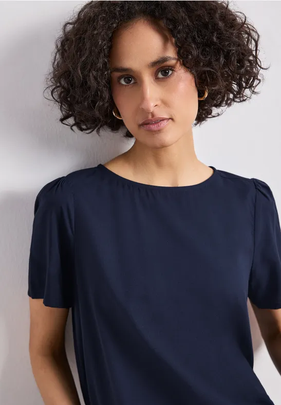 Een persoon met krullend haar draagt een marineblauw kort mouwtoppie voor een witte achtergrond.