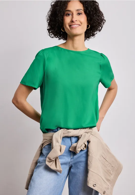 Person mit lockigen Haaren trägt grünes T-Shirt, Jeans und beige Jacke um die Taille, vor neutralem Hintergrund.