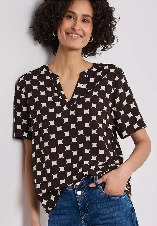 Frau trägt schwarz-weiße, kurzärmelige Bluse mit geometrischem Kreis- und Karo-Muster, teilweise in Jeans gesteckt.