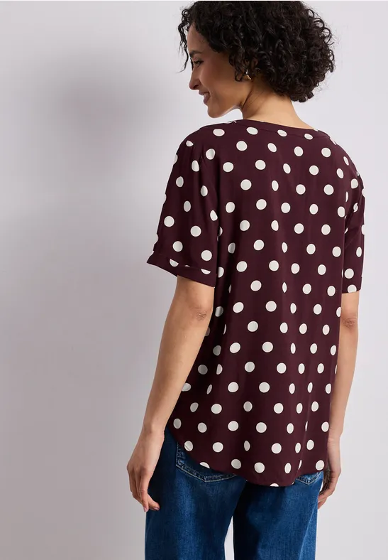 Femme de dos portant un t-shirt bordeaux à manches courtes, imprimé à pois blancs, et un jean.