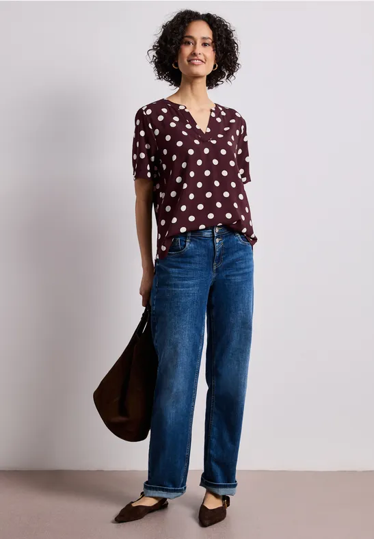 Femme en chemisier bordeaux à pois blancs, jean bleu, ballerines noires et sac marron.