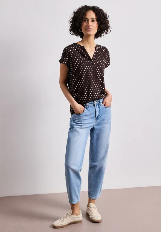 Une femme aux cheveux bouclés porte un t-shirt noir à pois, un jean bleu et des baskets blanches.
