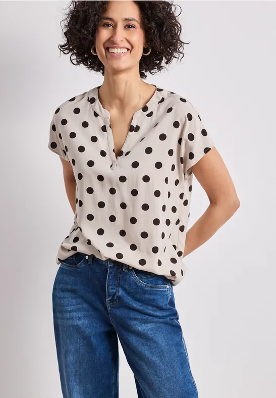 Femme en jean bleu et chemisier beige à pois noirs à manches courtes, mains derrière le dos.