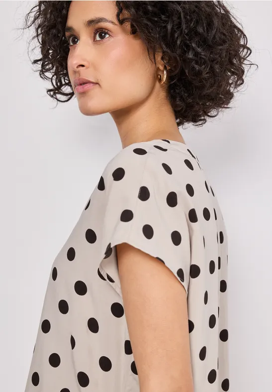 Femme de profil avec cheveux bouclés portant un chemisier beige à pois noirs.