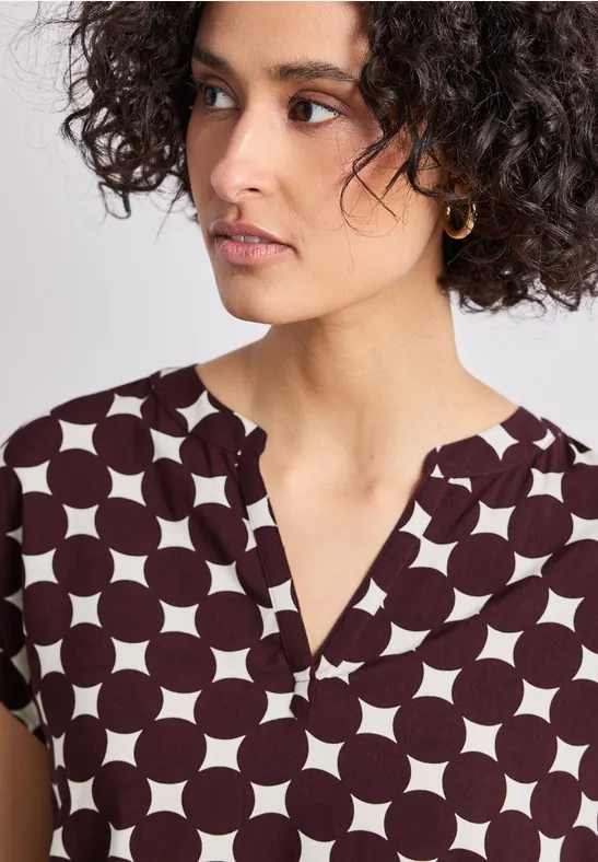 Portrait d'une femme aux cheveux bouclés, portant un haut blanc à gros pois brun foncé.