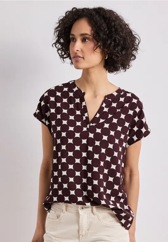 Femme aux cheveux bouclés porte un chemisier à manches courtes à motifs géométriques et un pantalon clair.