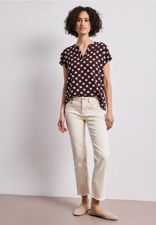 Une femme aux cheveux bouclés porte un chemisier noir à pois blancs, un pantalon clair et des mules.