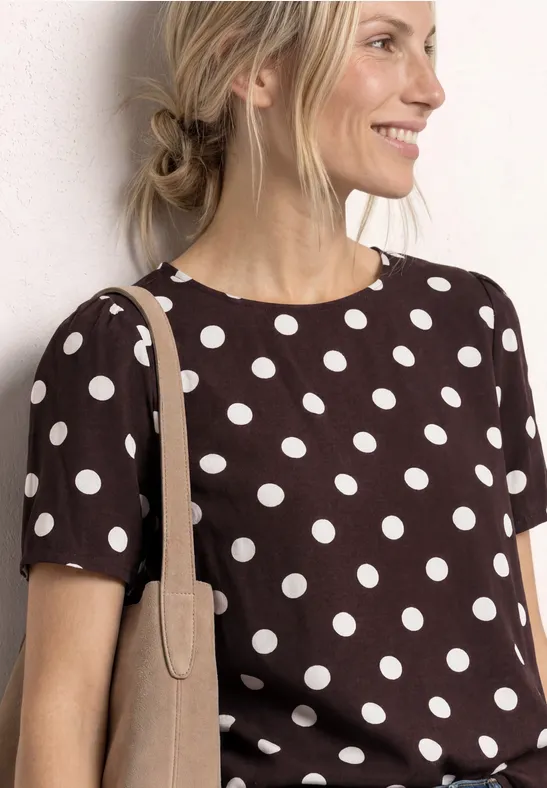 Femme en tee-shirt marron à pois blancs, sac beige à l'épaule, cheveux attachés en chignon lâche.
