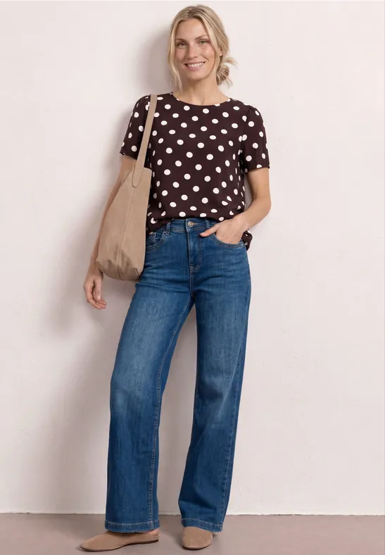 Femme en chemisier à pois, pantalon jean large bleu et sac porté épaule, posant décontractée.