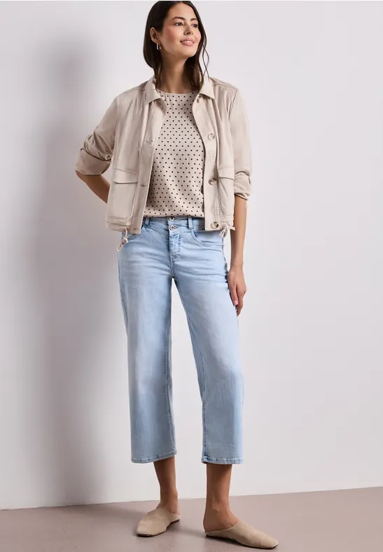 Vrouw draagt een beige jasje, een patroontrui, lichtblauwe cropped jeans en beige mules.