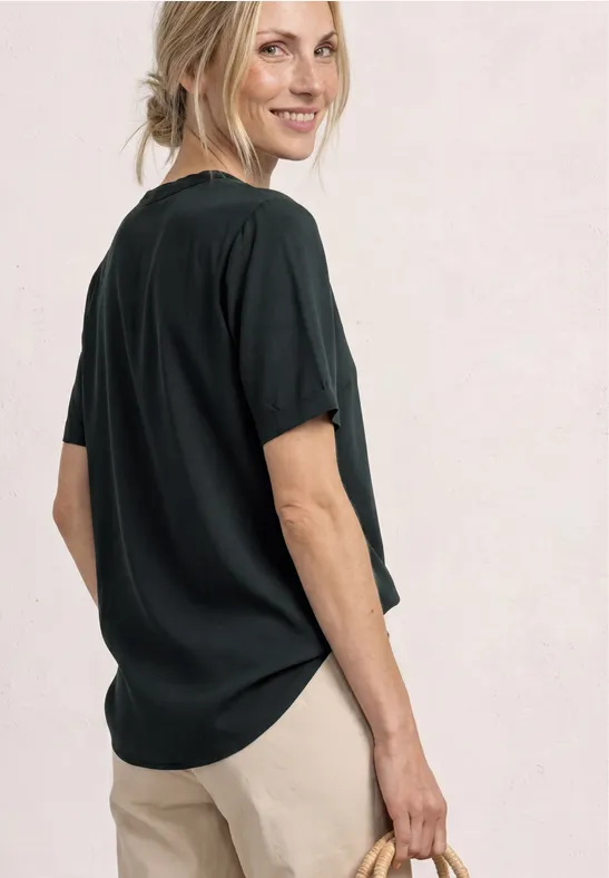 Frau im lässigen schwarzen T-Shirt und beigen Hosen, leicht seitlich, vor hellem Hintergrund.