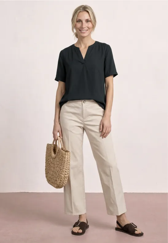 Frau in schwarzer Bluse, beiger Hose, schwarzen Sandalen und Korbtasche vor schlichtem Hintergrund.
