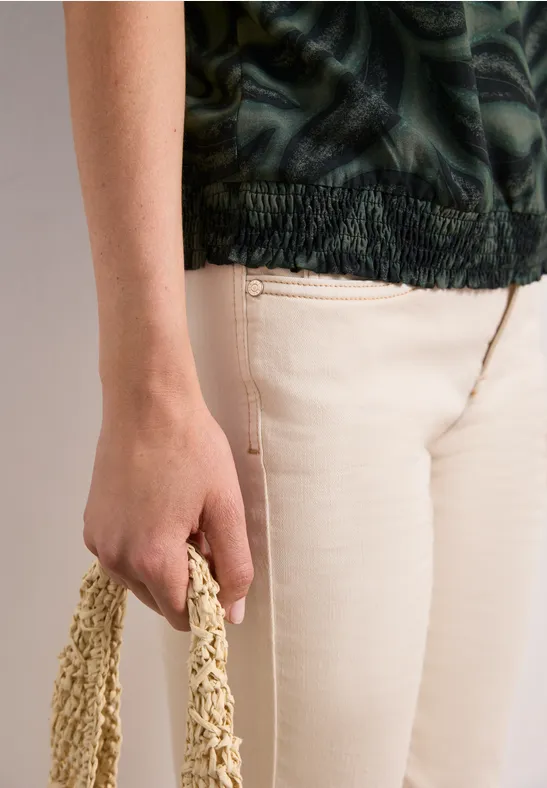 Person in beiger Hose und grünem Muster-Top hält eine geflochtene Tasche in der Hand.