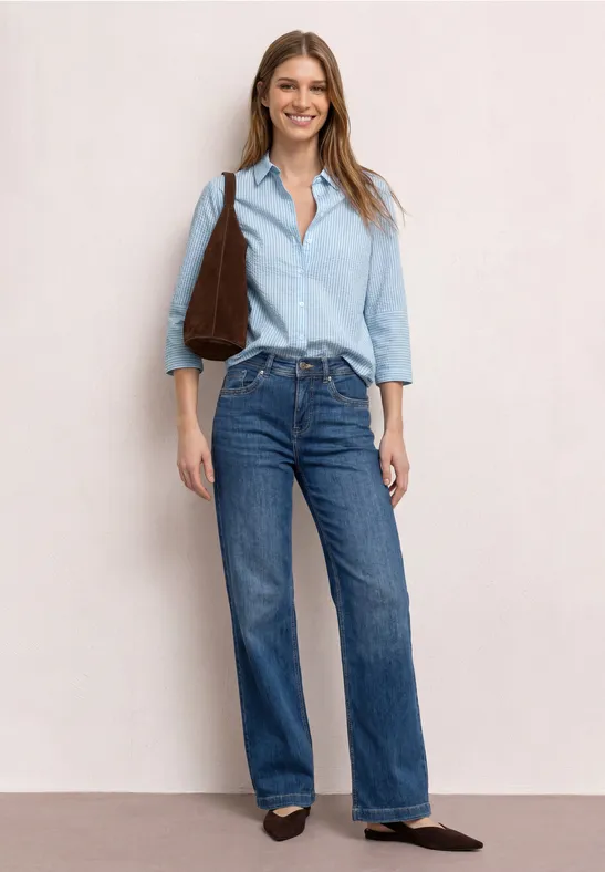 Vrouw in lichtblauw overhemd, blauwe jeans, bruine tas en schoenen voor neutrale achtergrond.