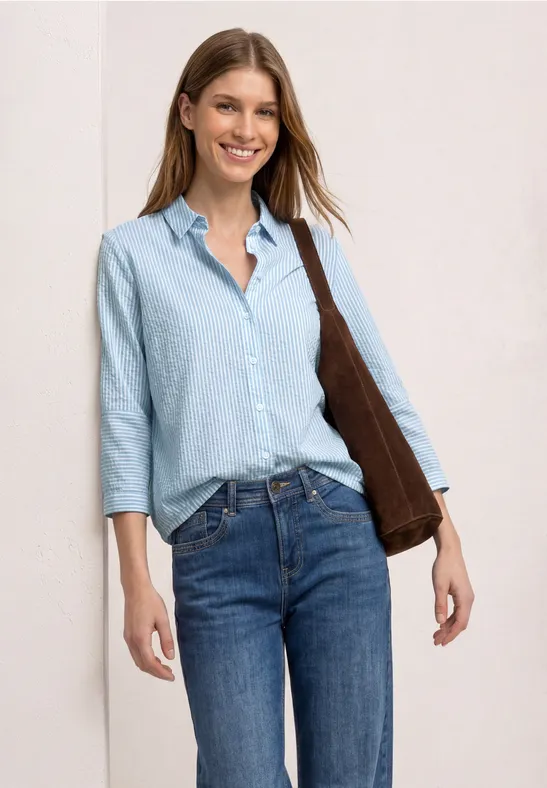 Vrouw met lichtblauw overhemd, driekwart mouwen, blauwe jeans en een bruine schoudertas.