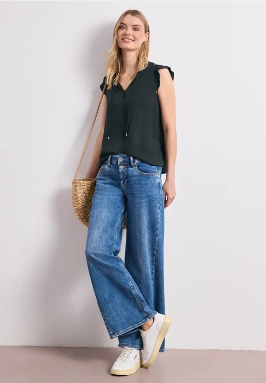 Frau in schwarzer ärmelloser Bluse, weiten Jeans, weißen Sneakern mit Strohtasche vor hellem Hintergrund.