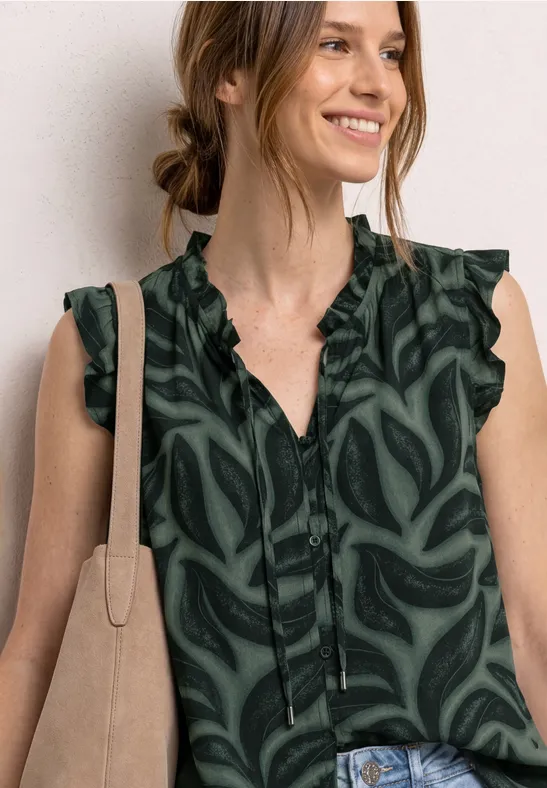 Vrouw in groene blouse met patroon, ruches en V-hals, met beige tas en spijkerbroek.