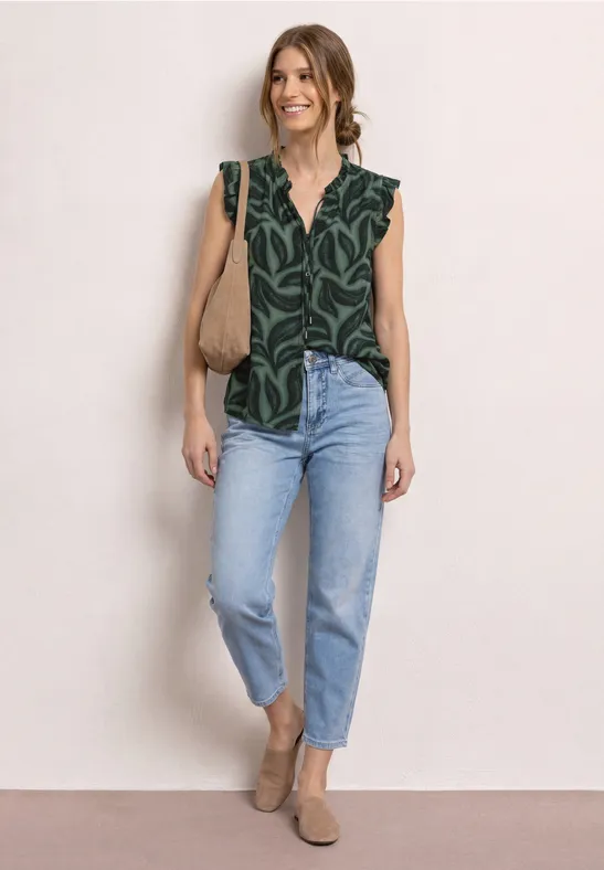 Vrouw in groen gemusterd topje, lichtblauwe jeans, bruine instappers en schoudertas, neutrale achtergrond.