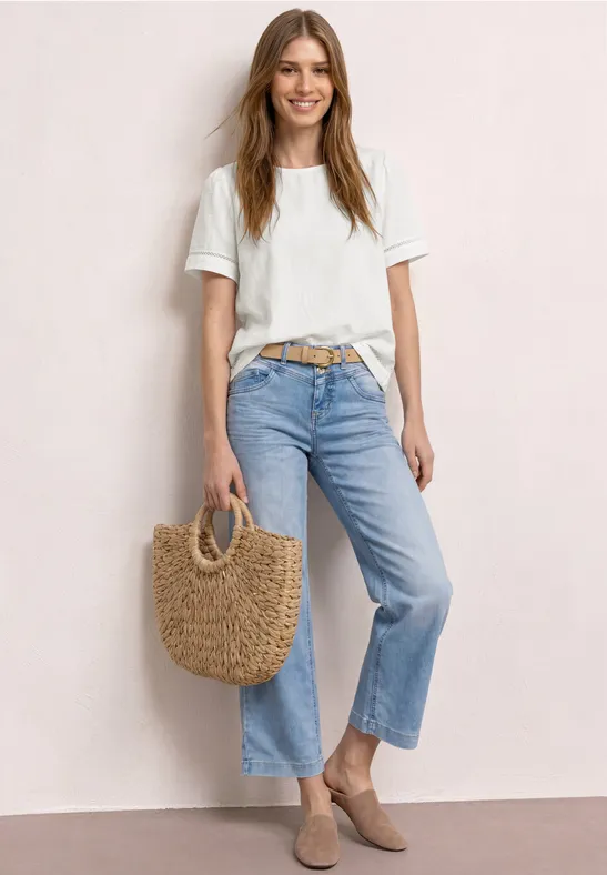 Vrouw in wit T-shirt, lichtblauwe jeans en beige loafers met een gevlochten strooien tas.