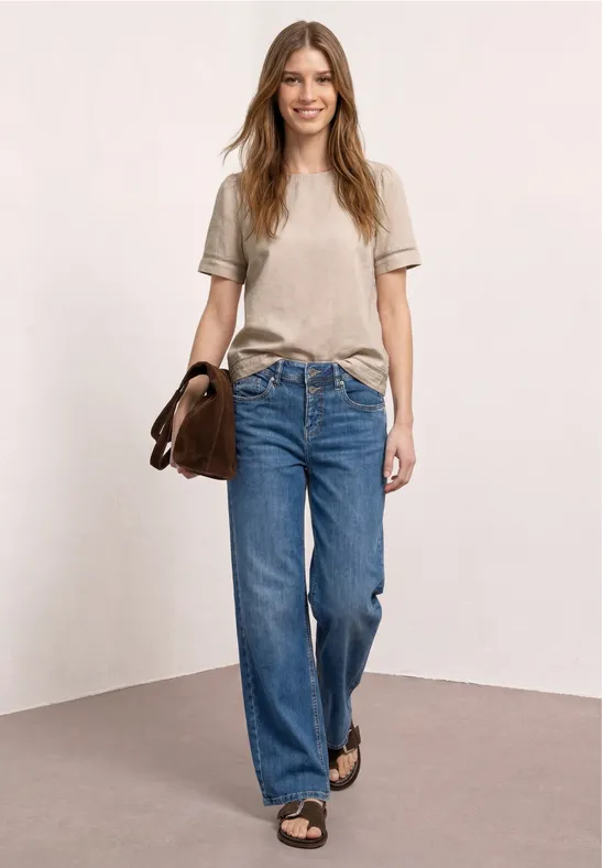 Vrouw in beige T-shirt, wijde spijkerbroek en sandalen met bruine tas voor een neutrale achtergrond.